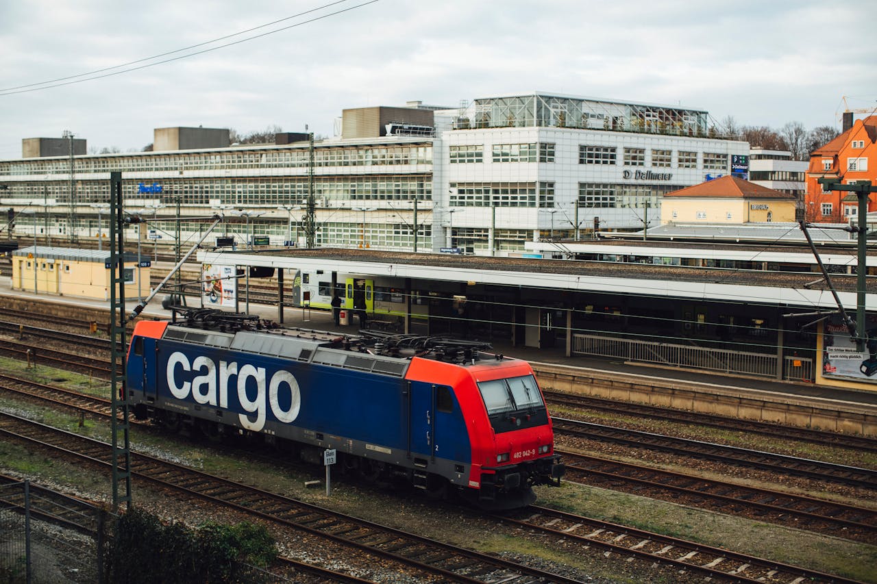 portfolio-06 Cargo train parked at a modern urban railway station on a cloudy day.